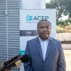 A man in a grey suit and white shirt is standing in front of a banner for ACEP, which includes text in French and Malagasy. He appears to be giving a media interview, as microphones and a smartphone are pointed toward him. The setting is outdoors, with trees and a wall in the background.