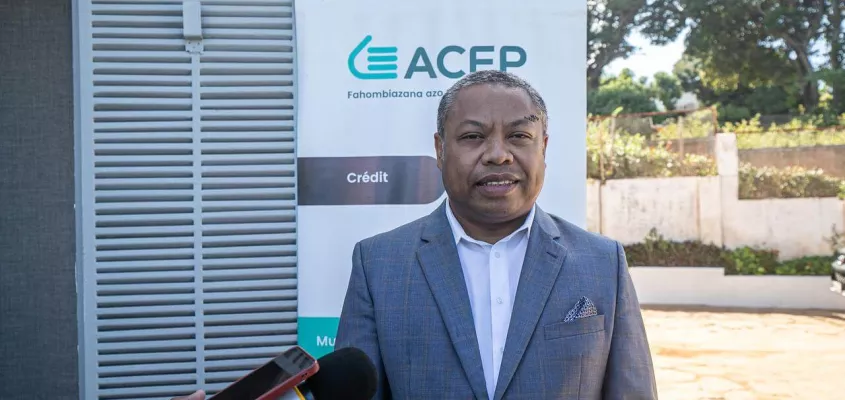 A man in a grey suit and white shirt is standing in front of a banner for ACEP, which includes text in French and Malagasy. He appears to be giving a media interview, as microphones and a smartphone are pointed toward him. The setting is outdoors, with trees and a wall in the background.